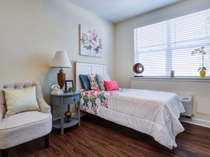A bright bedroom with a single bed covered in a white quilt with floral patterns and multiple pillows. Next to the bed is a small round gray nightstand with a lamp, a vintage telephone, and a small plant. A cushioned beige armchair with a light-colored pillow is positioned to the left. A large window with white blinds lets in natural light, and a floral painting hangs on the wall above the bed.