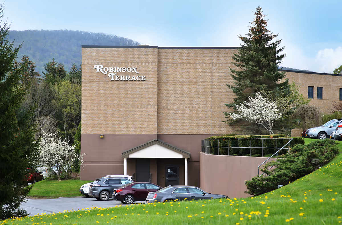 Exterior view of Robinson Terrace Rehabilitation and Nursing Center, a brick building with a small entrance canopy. Several cars are parked in front, surrounded by green grass, trees, and flowering bushes with a mountain visible in the background under a clear sky.