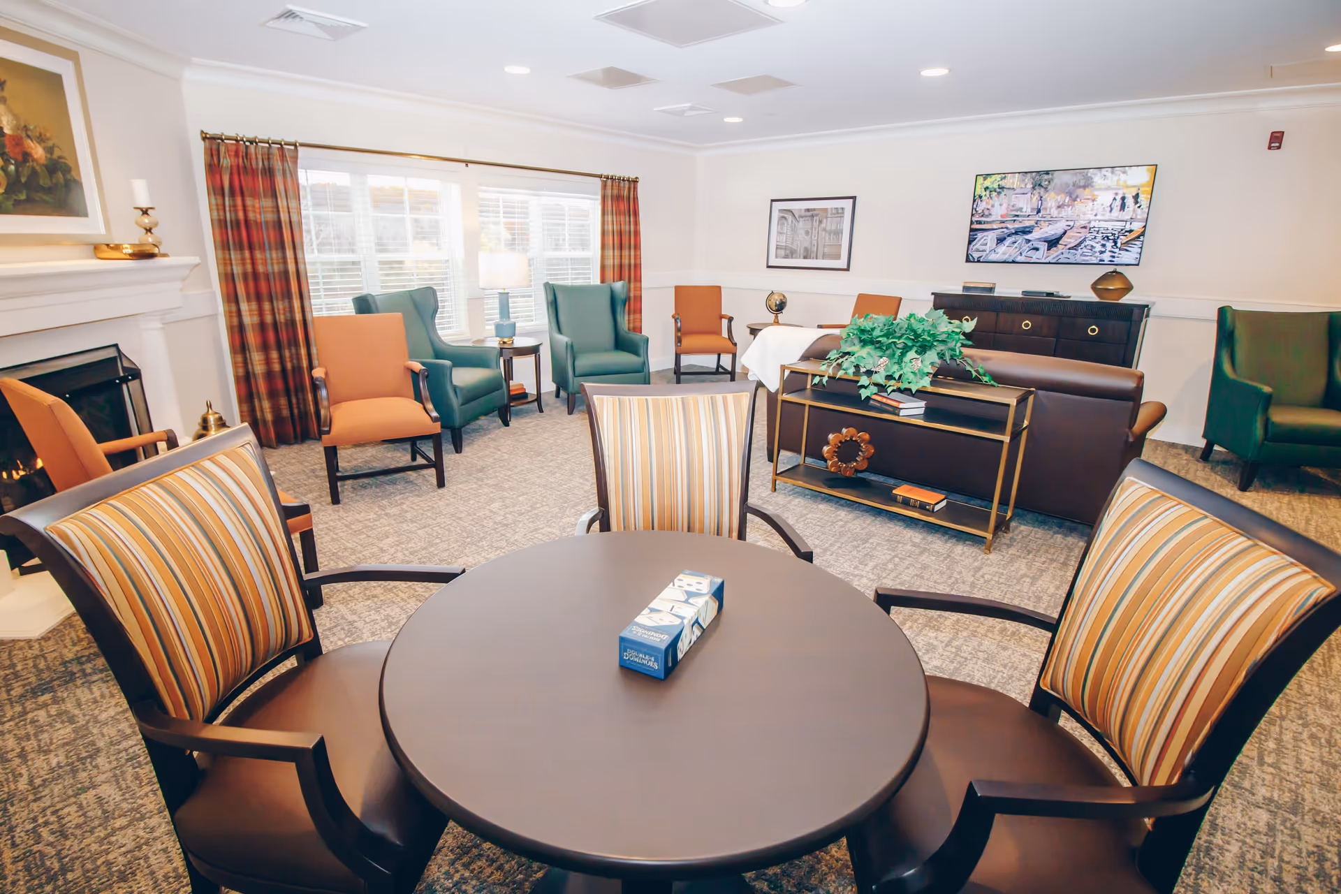 A bright and cozy senior living common area with a round table and four striped cushioned chairs in the foreground. Behind the table are several armchairs in green and orange colors arranged near a window with plaid curtains. The room features a fireplace on the left, a brown leather sofa with a console table behind it holding books and a plant, and framed artwork on the walls.