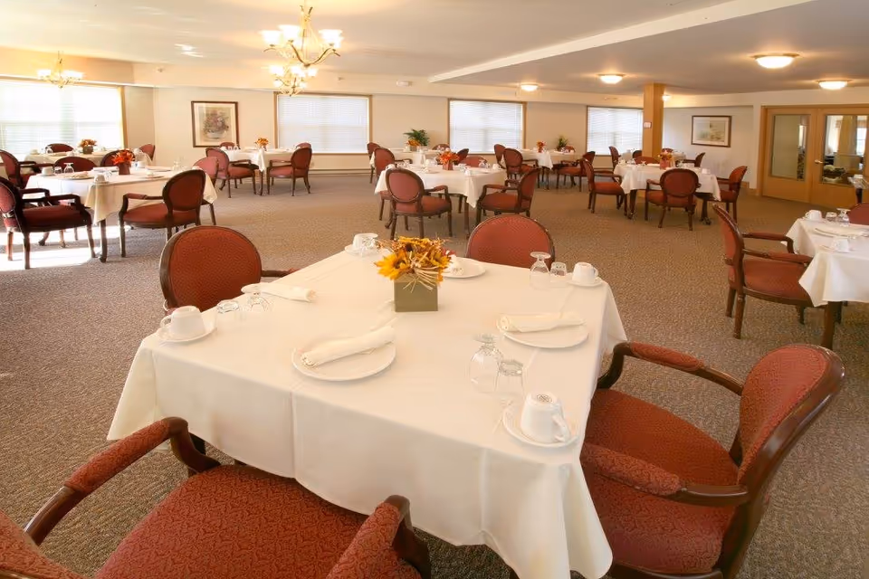 A spacious dining room with multiple tables covered in white tablecloths, each set with plates, cups, napkins, and glasses. The chairs have red upholstery and wooden frames. The room is well-lit with ceiling lights and chandeliers, and there are large windows with blinds along the walls. Some framed artwork and plants are visible in the background.
