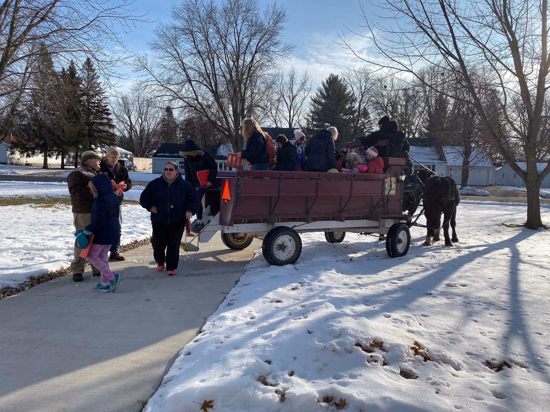 A group of people, including children and adults, are gathered around and sitting in a horse-drawn wagon on a snowy day. The wagon is on a paved path with bare trees and houses visible in the background under a clear blue sky.