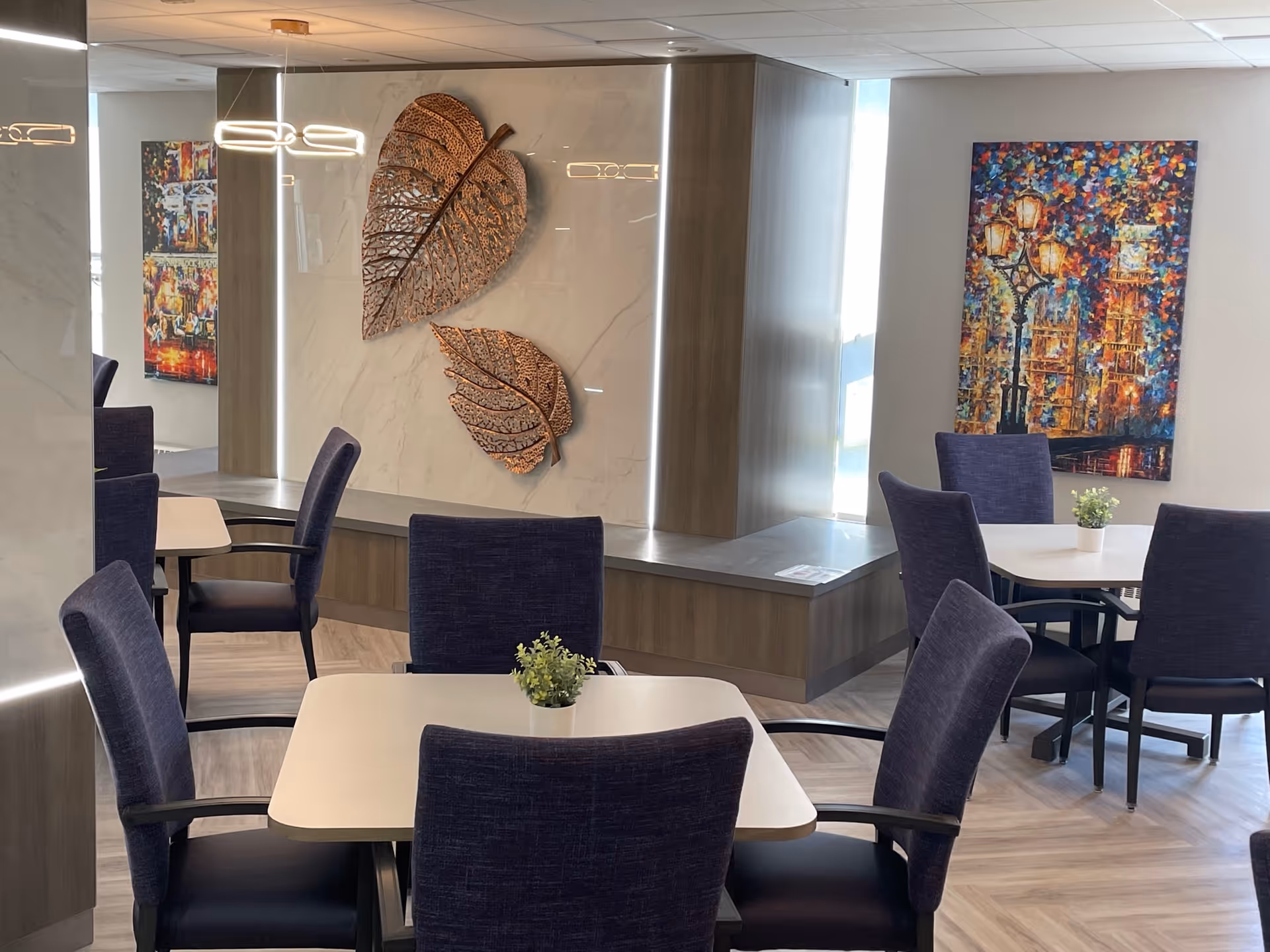 A modern dining area with several square tables and dark upholstered chairs. Each table has a small green plant centerpiece. The walls feature decorative elements including two large metallic leaf sculptures and a colorful painting of a streetlamp and buildings. The floor has a light wood pattern and the space is well-lit with natural light from a window.