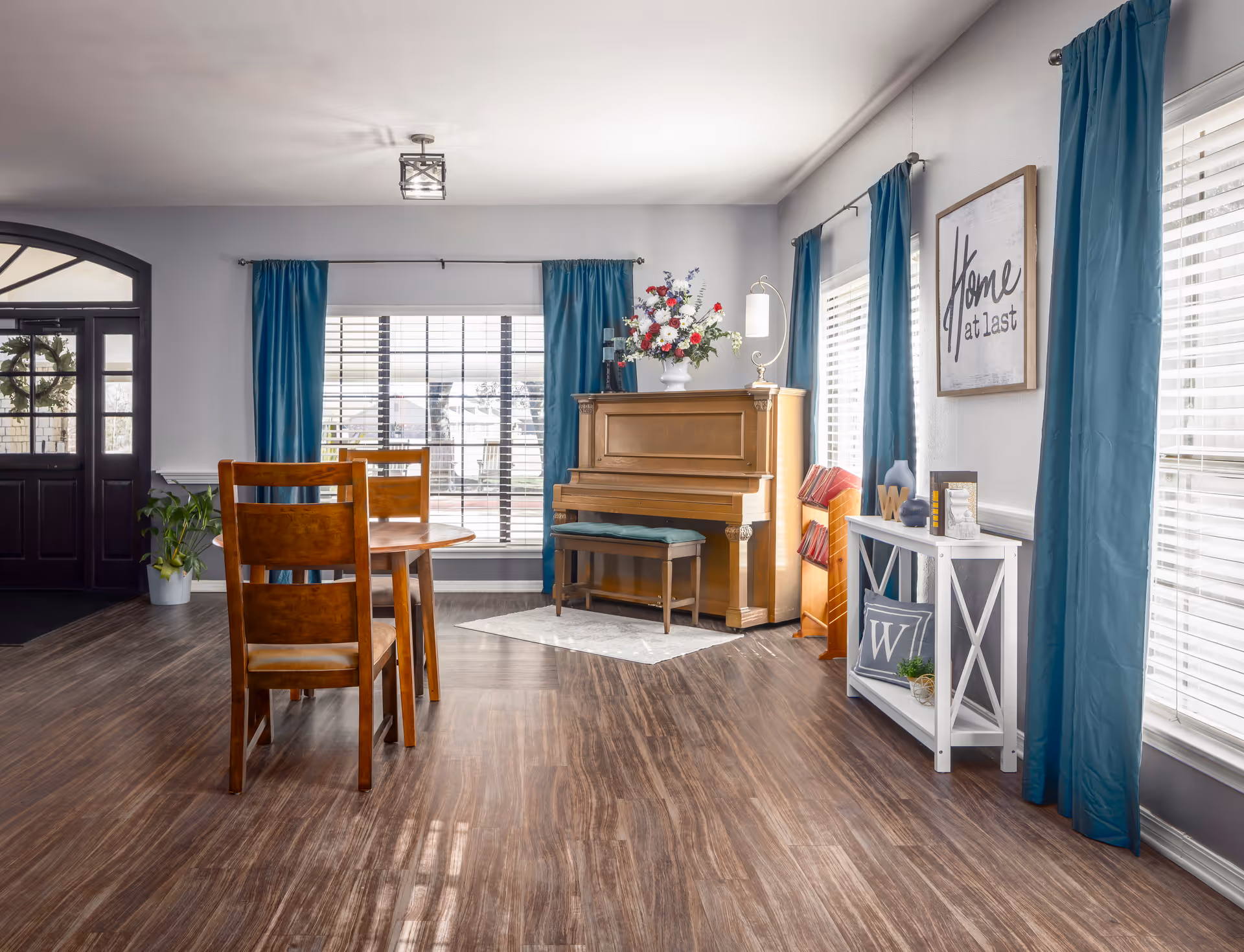 A bright and cozy living room area featuring a wooden piano with a floral arrangement on top, a small wooden dining table with chairs, large windows with blue curtains, and a white shelf with decorative items. A framed sign on the wall reads 'Home at last'.
