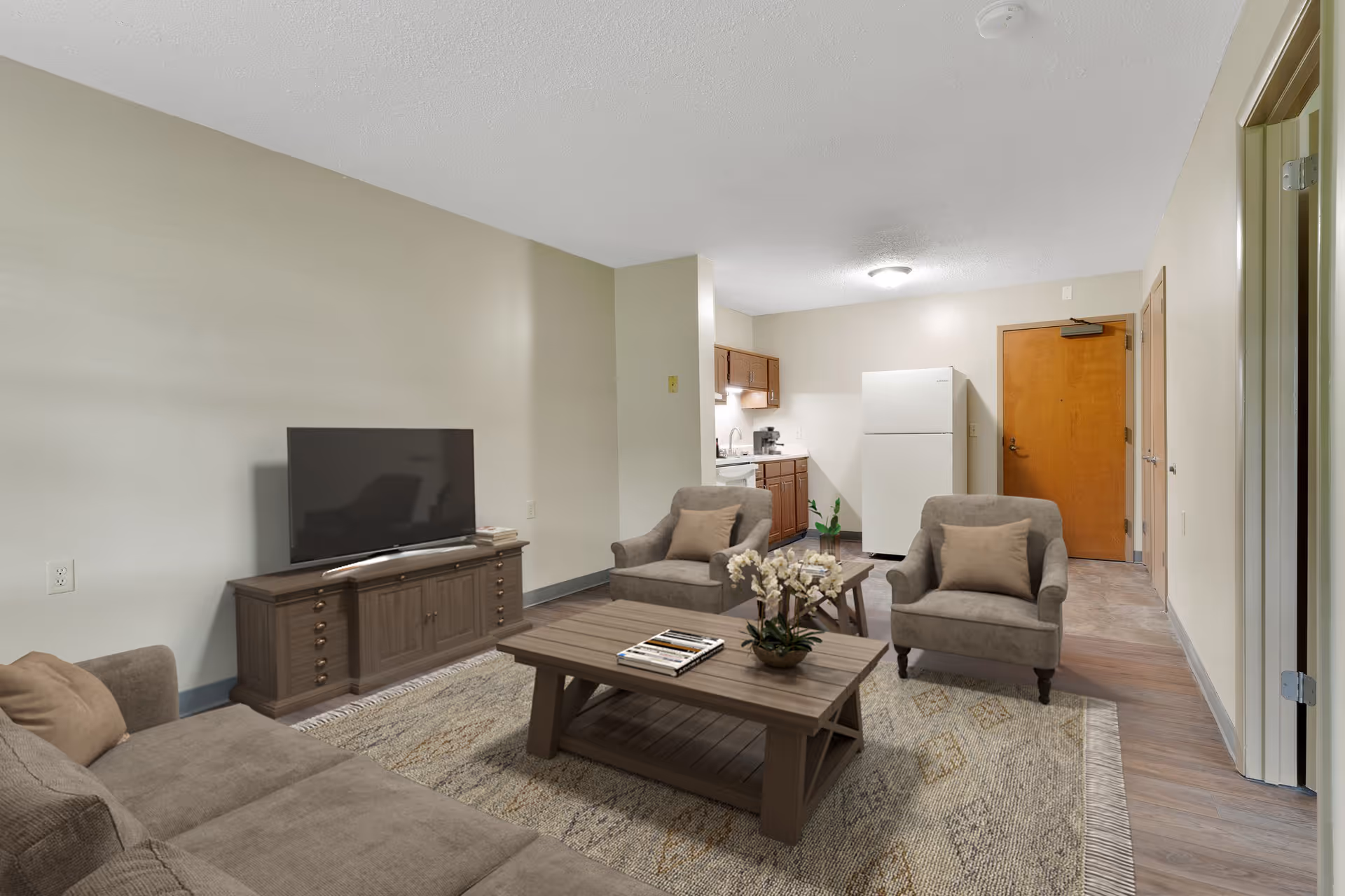 A cozy living room area with a beige sofa, two armchairs with cushions, a wooden coffee table with a flower arrangement and magazines, a TV on a wooden stand, and a small kitchen area with wooden cabinets, a refrigerator, and a coffee maker. The room has light-colored walls and wood flooring.