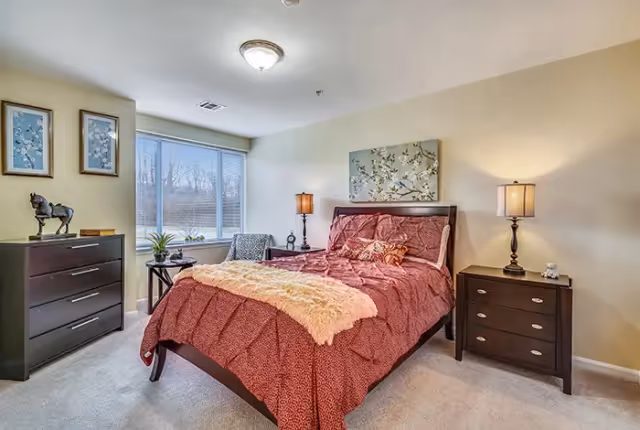 A cozy bedroom with a large window letting in natural light. The room features a bed with a red patterned comforter and a beige throw blanket, two dark wooden nightstands each with a lamp, a dark wooden dresser with decorative items including a horse figurine, and two framed pictures on the wall.