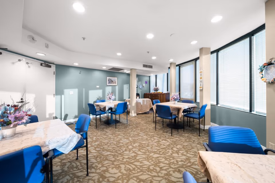 A bright dining room with several tables covered in light-colored tablecloths and decorated with floral centerpieces. Blue chairs surround the tables. Large windows with blinds allow natural light to fill the room. The walls are painted in a soft green color, and the carpet has a patterned design. There is a wooden cabinet against the far wall with additional floral arrangements and decorations.