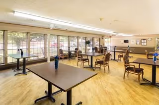 Spacious dining room with multiple tables and chairs arranged near large windows in a senior living facility.