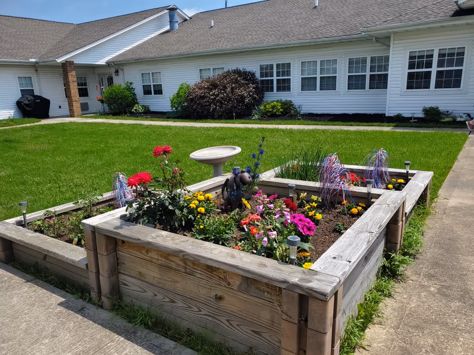 Raised wooden garden beds filled with colorful flowers and decorative items, situated on a concrete pathway with a green lawn and white building in the background under a clear sky.