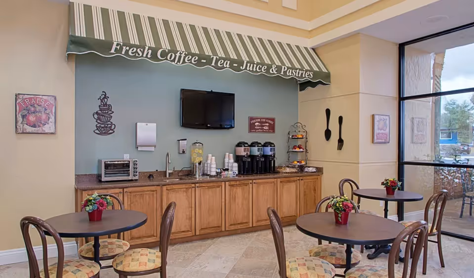 A cozy dining area with round tables and chairs featuring patterned cushions. A countertop with wooden cabinets below holds coffee dispensers, a toaster oven, a water dispenser with lemon slices, and disposable cups. Above the counter is a wall-mounted TV and a green striped awning with the text 'Fresh Coffee - Tea - Juice & Pastries'. The walls are decorated with coffee-themed art and large utensils. Large windows allow natural light into the space.