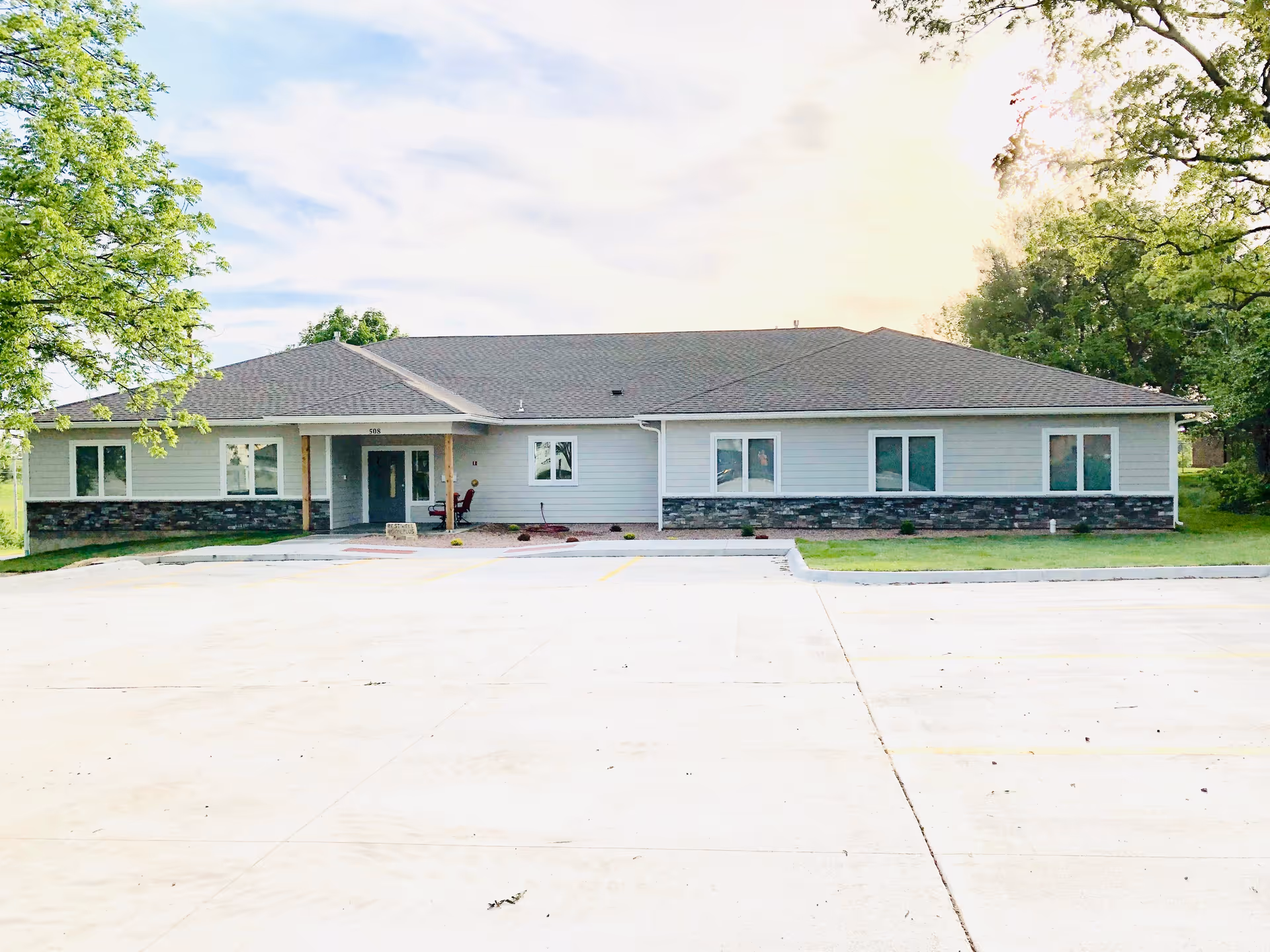 Single-story building with gray siding and stone accents, surrounded by green trees and a large empty concrete parking lot in front under a partly cloudy sky.