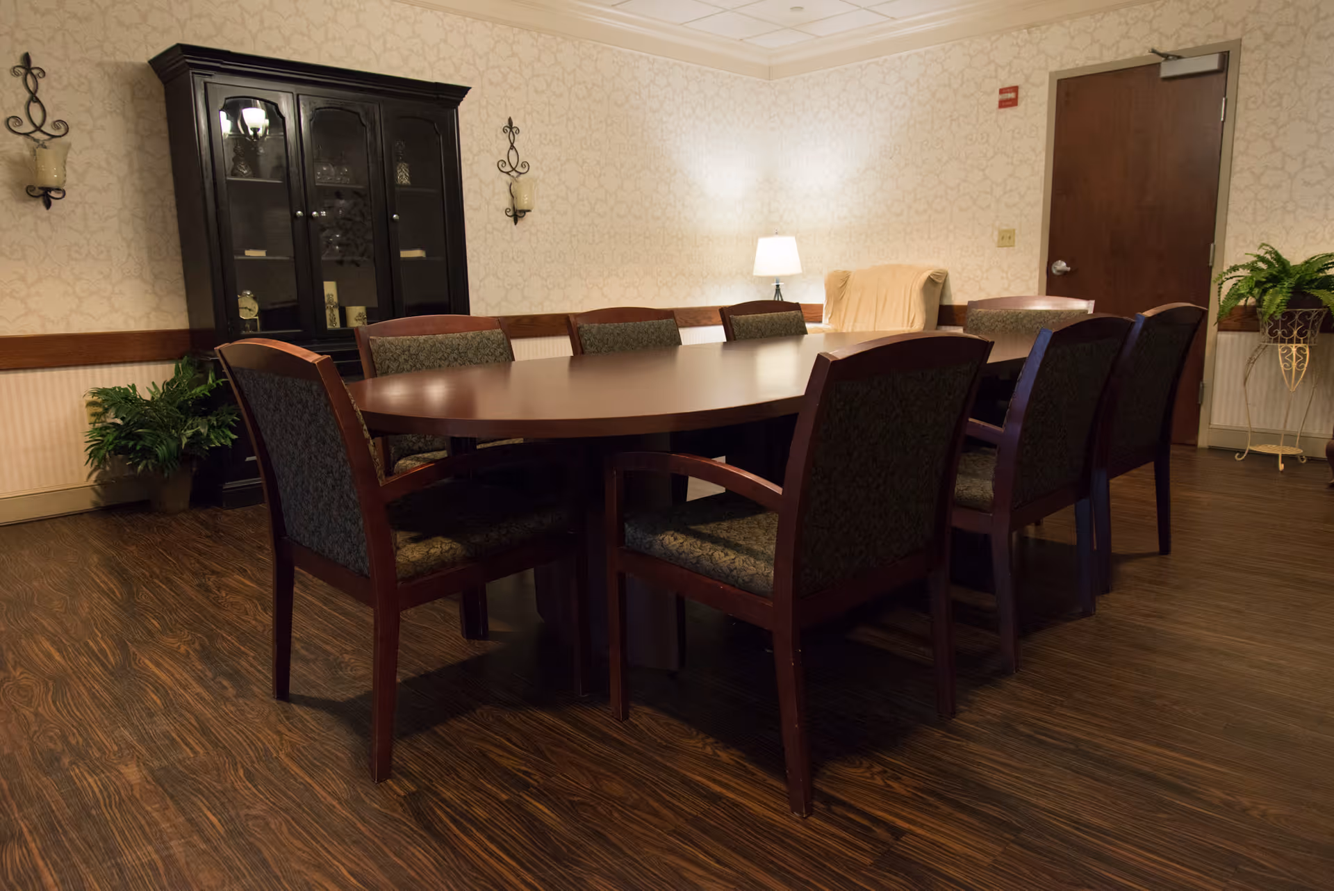 A well-lit room with a large oval wooden table surrounded by eight cushioned chairs. The room has wood flooring, patterned wallpaper, a dark wooden cabinet with glass doors, two wall-mounted candle holders, a potted plant, a floor lamp, and a closed wooden door.
