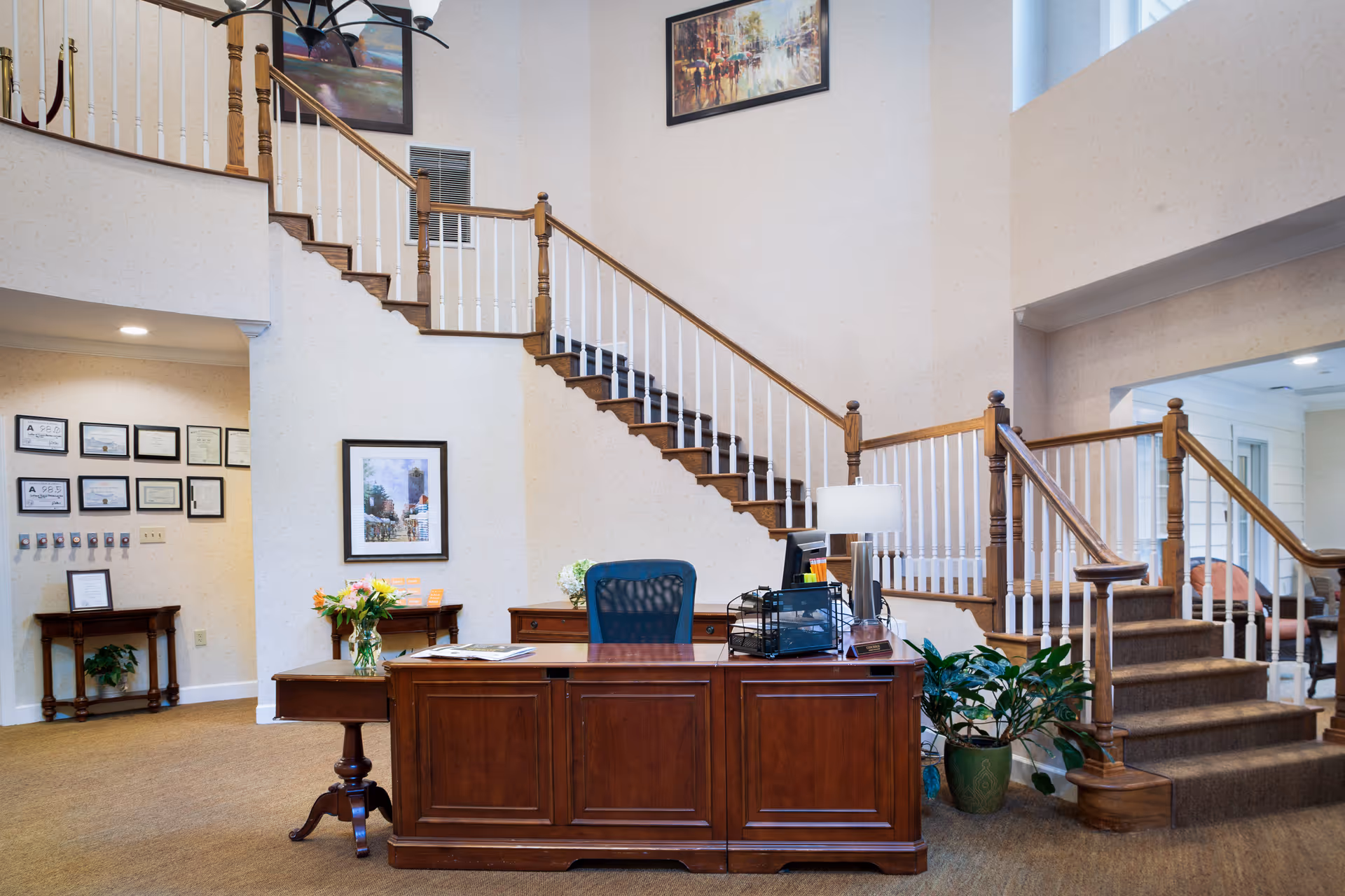 Reception area with a wooden desk and office chair in front of a staircase with wooden handrails and white balusters. The walls are decorated with framed pictures and certificates. There are plants and a table with flowers near the desk. The space is well-lit with a high ceiling and a chandelier.