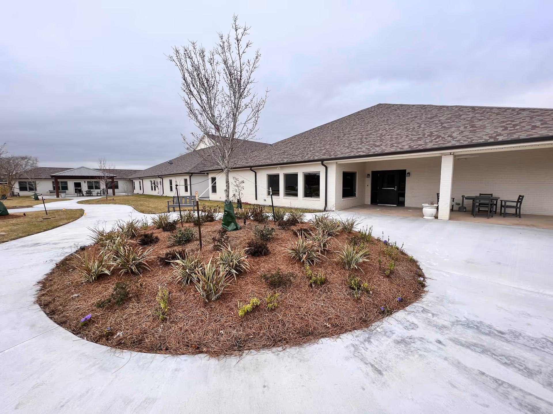Outdoor view of a senior living facility with a circular landscaped area featuring small plants and a young tree in the center. The building has a light-colored exterior with a covered patio area furnished with tables and chairs. The sky is overcast.