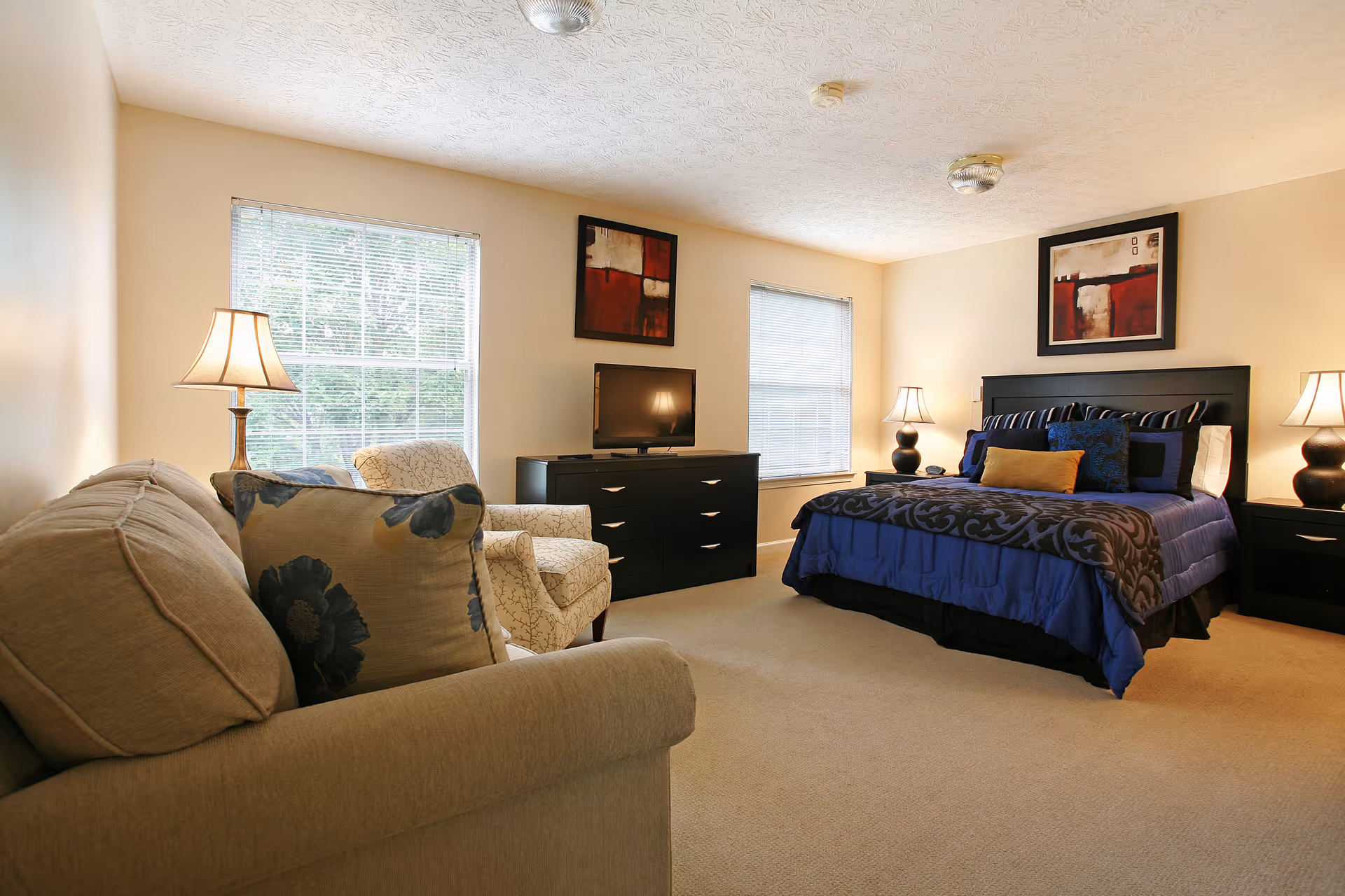 A well-lit bedroom featuring a large bed with blue and black bedding, two bedside tables with lamps, a black dresser with a flat-screen TV, a beige armchair, and a beige sofa with floral pillows. Two windows with blinds allow natural light into the room, and abstract artwork hangs above the bed and dresser.