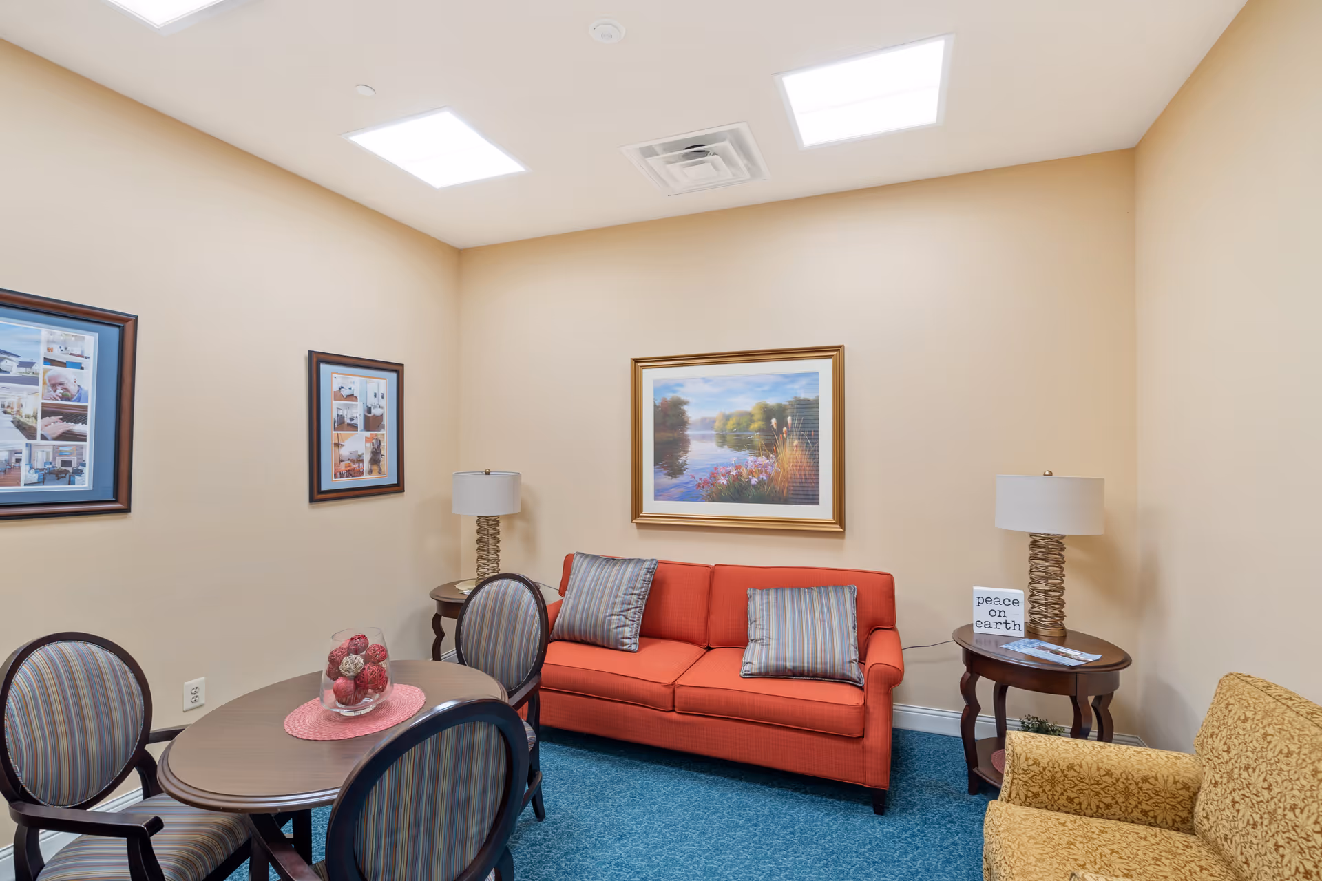 A cozy sitting area in a memory care community featuring a red sofa with two striped pillows, a yellow armchair, a round wooden table with four striped chairs, two side tables with lamps, and framed artwork on beige walls.