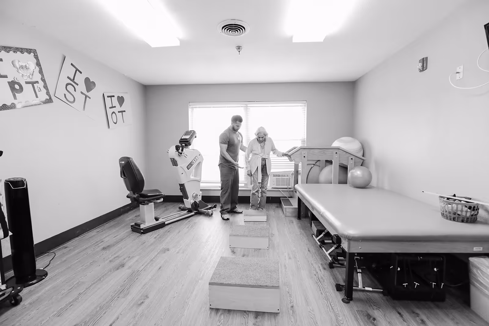 A physical therapy room with wooden flooring and exercise equipment. An elderly woman is stepping on wooden blocks while being assisted by a male therapist. The room has a large padded therapy table, exercise balls, and a stationary exercise bike. Signs on the wall say 'PT', 'I ♥ ST', and 'I ♥ OT'.