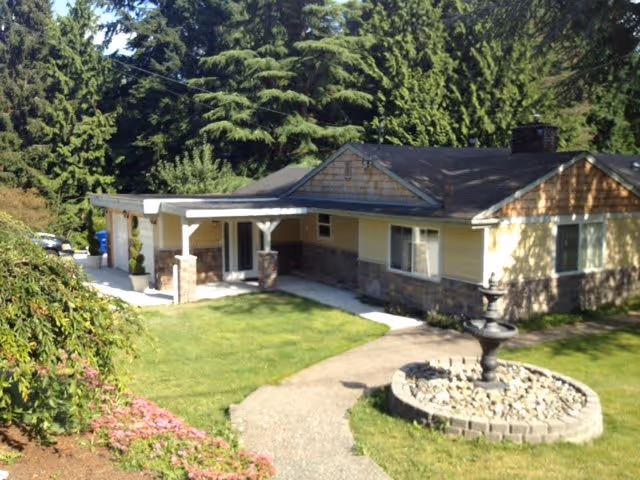 Single-story residential building with a covered entrance supported by white columns, surrounded by green grass and trees. A curved concrete pathway leads to a circular stone-bordered fountain in the front yard.