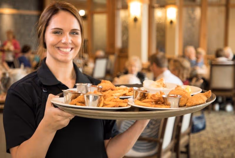 A smiling waitress holding a large tray with plates of fried food and dipping sauces in a busy dining room with people seated at tables in the background.