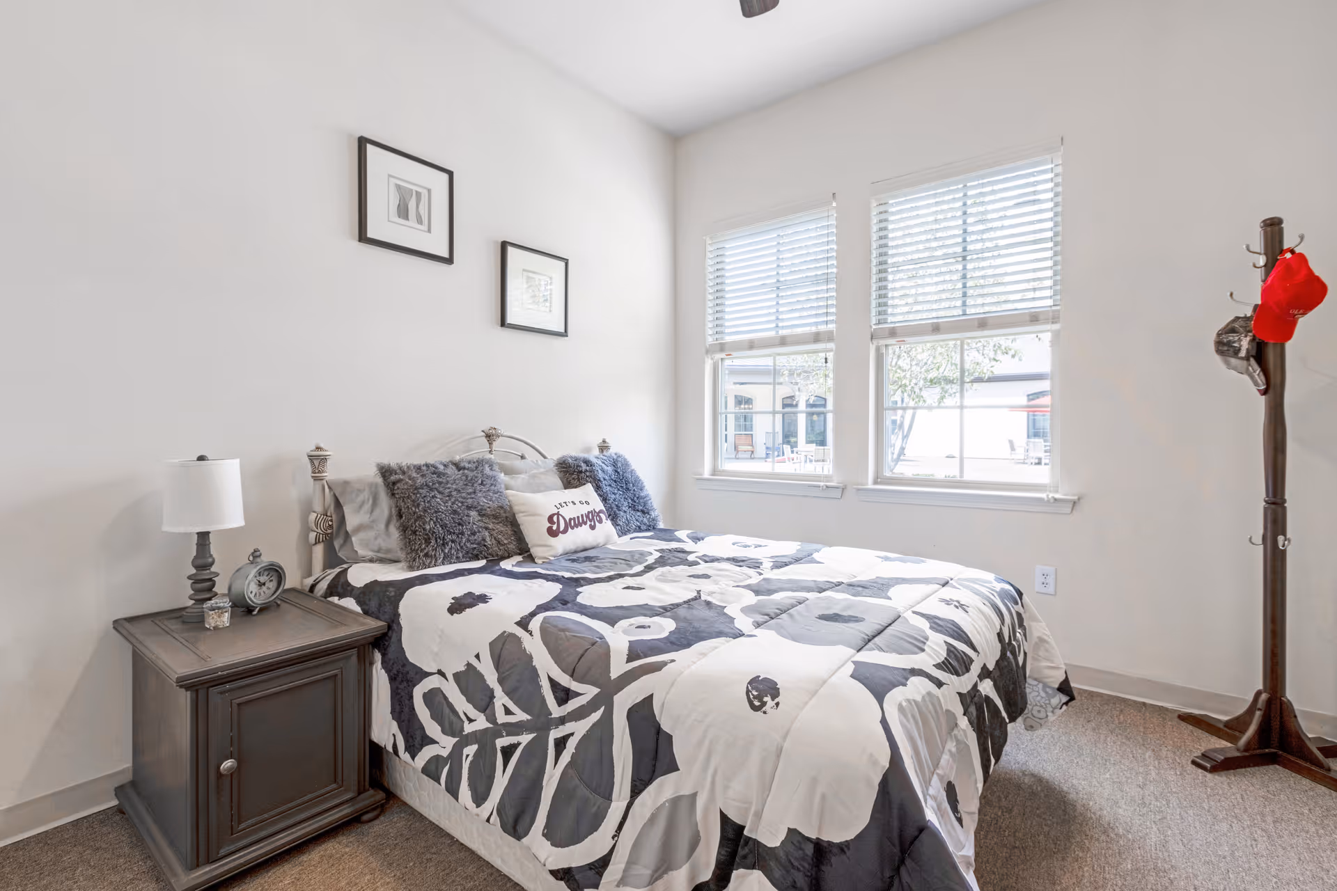 A bright bedroom with a large bed covered in a black and white floral patterned comforter. The bed has two gray fluffy pillows and a decorative pillow with the word 'Dawgs'. There is a wooden nightstand with a lamp and an alarm clock on the left side of the bed. Two framed pictures hang on the white wall above the bed. Two windows with white blinds let in natural light. A wooden coat rack with hats and a red cap stands in the corner on the right.