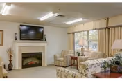 A cozy living room area with a fireplace beneath a mounted flat-screen TV. The room features a beige carpet, two striped armchairs, a floral patterned sofa, a wooden side table with a lamp, and a large window with beige curtains allowing natural light to enter.
