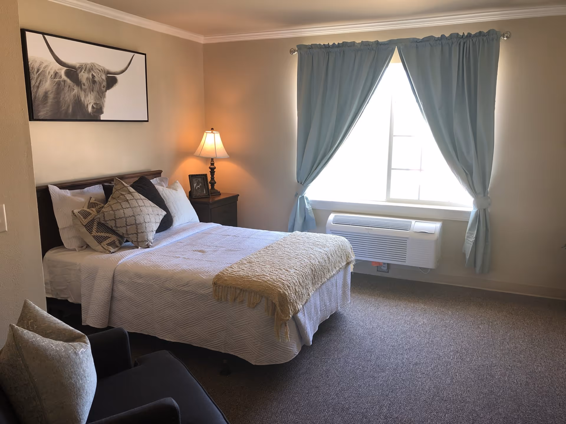 A bedroom with a neatly made bed featuring white bedding and multiple decorative pillows. There is a nightstand with a lamp and a framed photo next to the bed. A large window with light blue curtains allows natural light into the room. A framed picture of a highland cow hangs on the wall above the bed. A cushioned chair with a pillow is partially visible in the foreground.