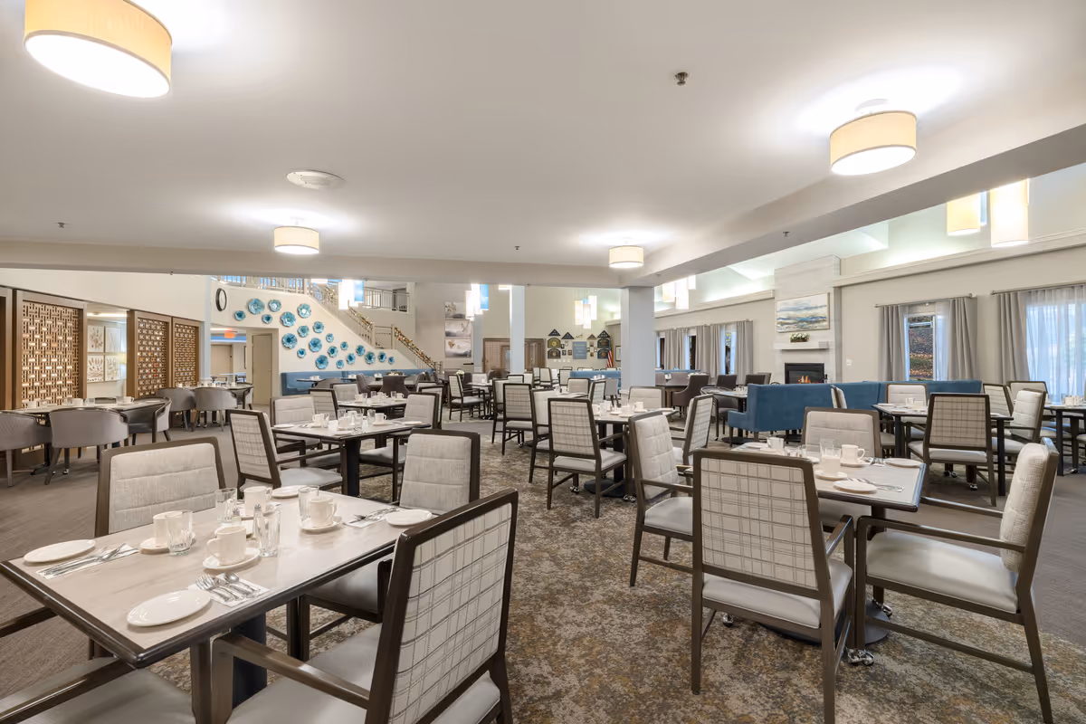 A spacious dining room in a senior living facility with multiple tables and chairs arranged neatly. Each table is set with white plates, cups, and silverware. The room features soft lighting from ceiling fixtures, a carpeted floor, and large windows with curtains. There is a fireplace with artwork above it and decorative blue plates on the wall near a staircase in the background.