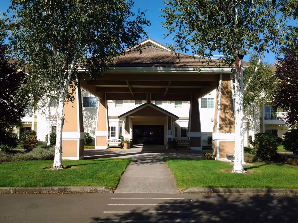Entrance of a retirement community building with a covered driveway supported by large pillars, flanked by green lawns and trees under a clear blue sky.