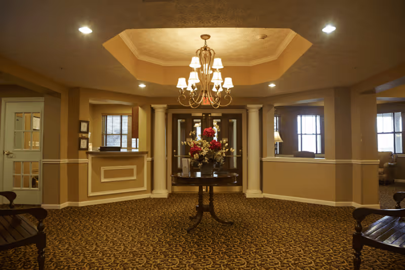 Lobby interior with a central round table topped with a floral arrangement, chandelier, patterned carpet, and seating areas.