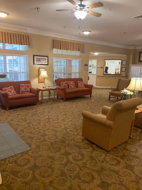 A cozy assisted living facility common area with patterned carpet, two red sofas with decorative pillows, a beige armchair, side tables with lamps, and windows with striped valances. In the background, there is a reception desk area with a person behind the counter.
