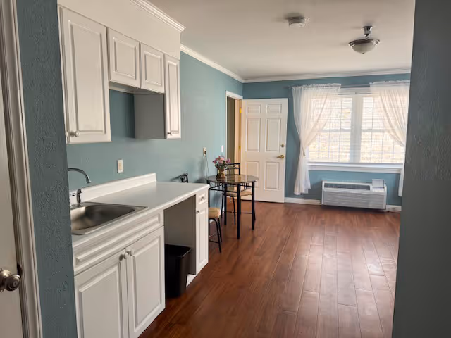 Interior view of a room in Roxboro Assisted Living showing a small kitchenette with white cabinets and a sink on the left, a small table with two chairs in the middle, a window with sheer white curtains, and a heating/cooling unit below the window. The walls are painted light blue and the floor is wood.