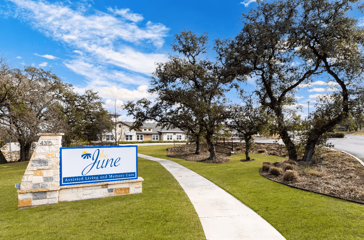 Outdoor view of a senior living facility with a stone sign that reads 'The June Assisted Living and Memory Care' next to a curved concrete pathway, surrounded by green grass, trees, and a clear blue sky with some clouds.