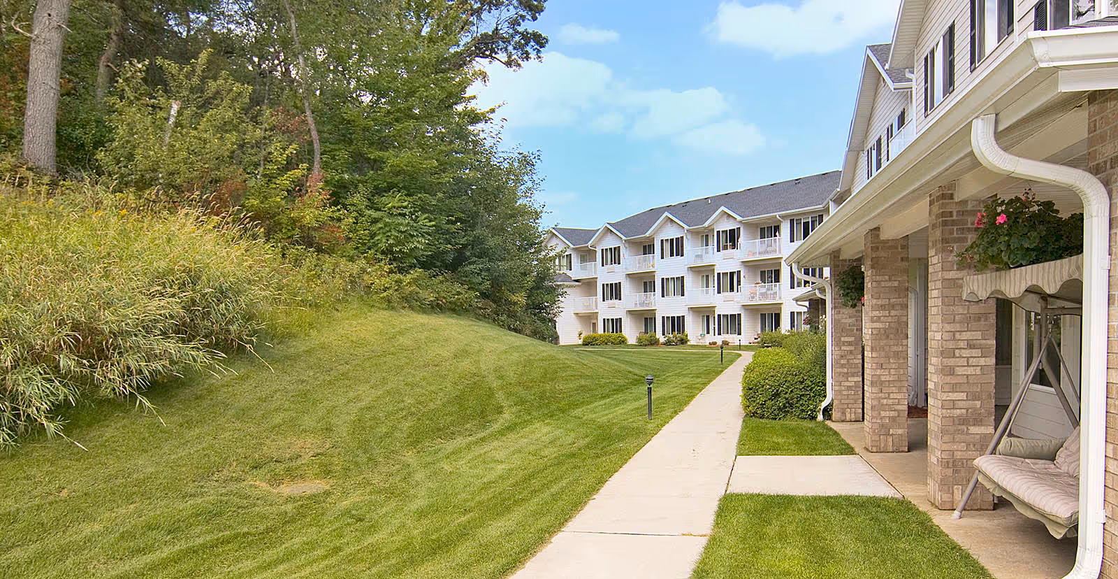 A paved walkway runs alongside a grassy lawn with bushes and trees on one side and a multi-story residential building on the other. The building has balconies and a covered porch area with a cushioned swing. The sky is partly cloudy.