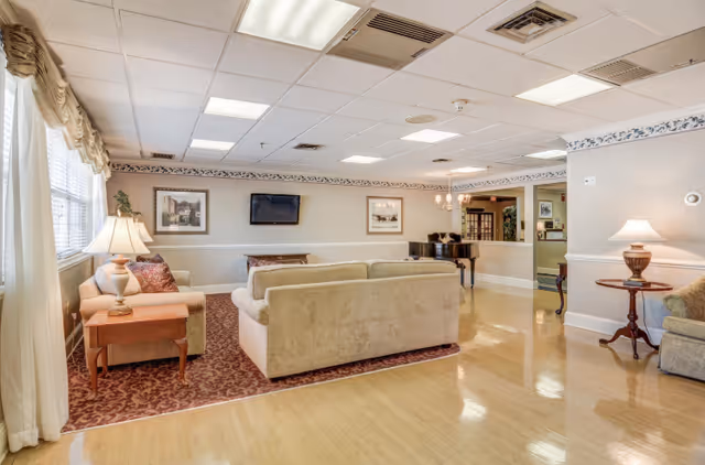 Spacious common lounge with sofas arranged on a rug facing a wall-mounted TV, side tables and lamps, and a piano in a senior living facility.