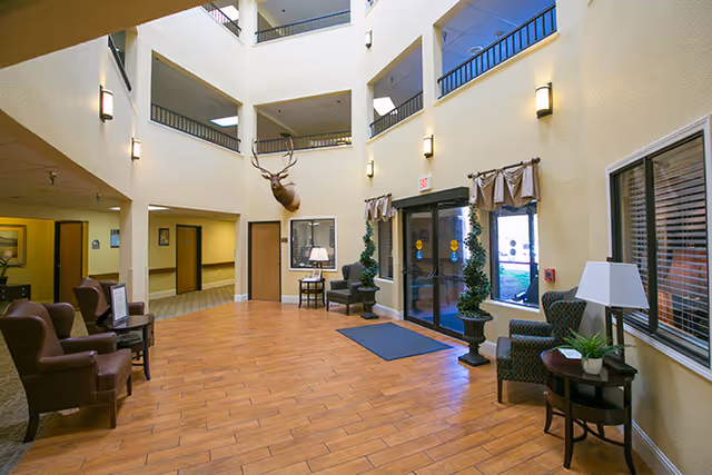 Interior view of a senior living facility lobby with high ceilings and multiple floors visible. The space features wooden flooring, several armchairs arranged along the walls, small tables with lamps and plants, and a mounted deer head on the wall. Large glass doors lead outside, flanked by decorative greenery.