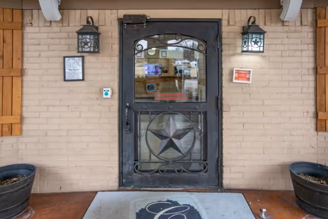 Entrance door of Carrollton Health & Rehabilitation Center with a decorative metal door featuring a star design. The door is flanked by two wall-mounted lantern-style lights and wooden shutters on the brick wall. There are two large planters on either side of the entrance and a welcome mat with a decorative letter C in front of the door.