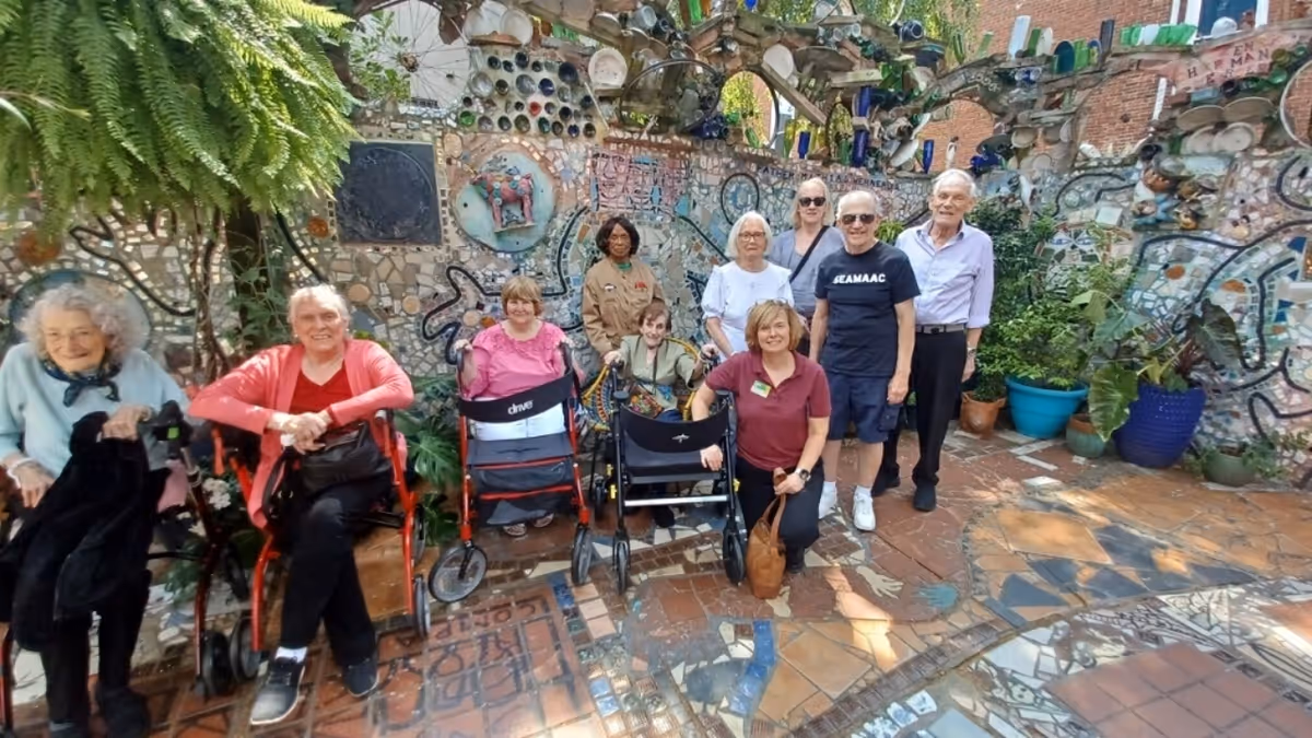 A group of elderly people and caregivers posing together in an outdoor mosaic garden area with colorful tiled walls and potted plants.