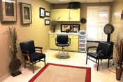 Small salon-style room with a central styling chair in front of yellow cabinets, two guest chairs, a hooded hair dryer, and decorative plants and wall art.