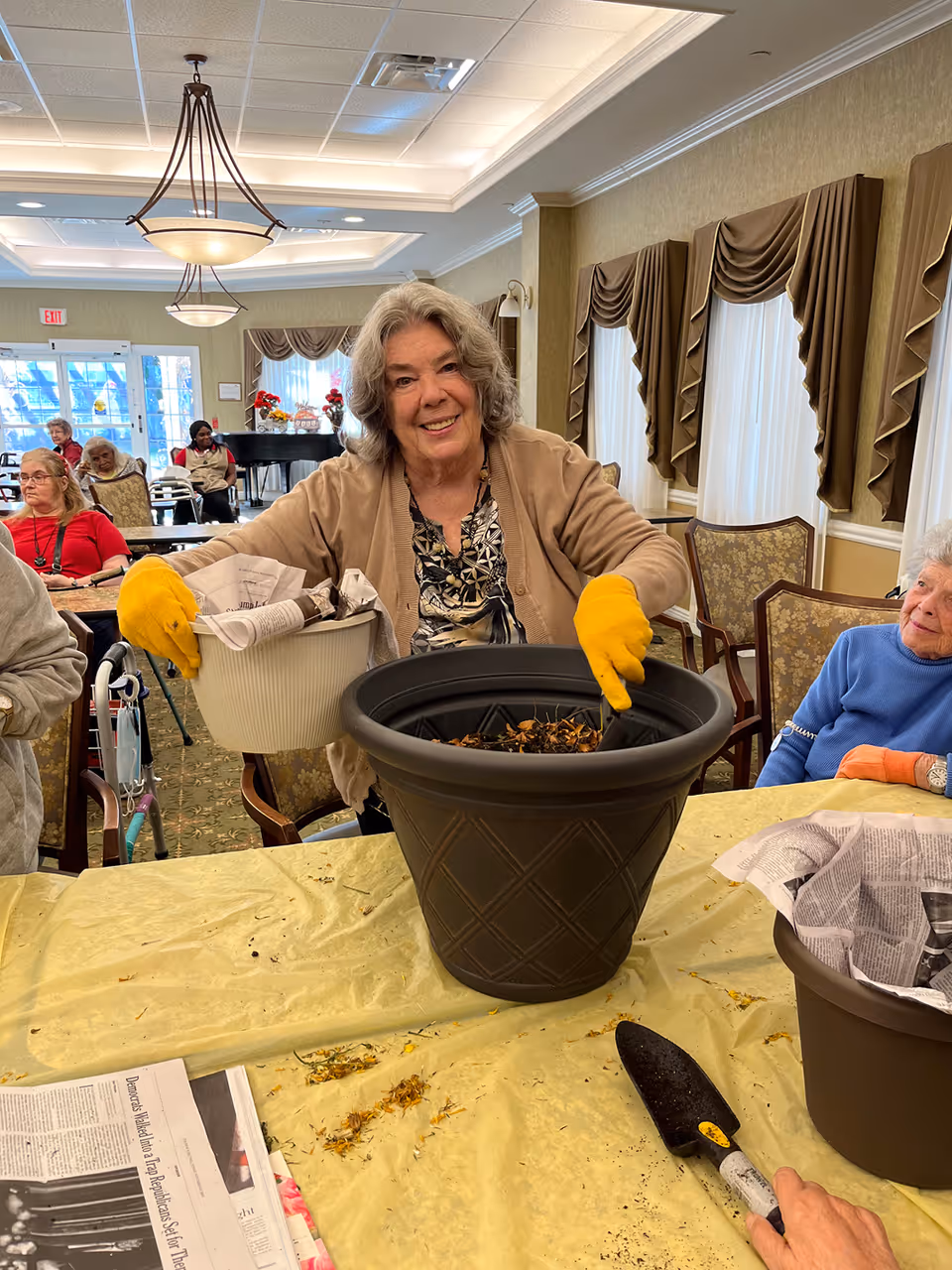 An elderly woman wearing yellow gloves is smiling while planting flowers in a large black pot inside a well-lit room with several other elderly people seated around tables. The room has beige walls, large windows with brown curtains, and a chandelier hanging from the ceiling.