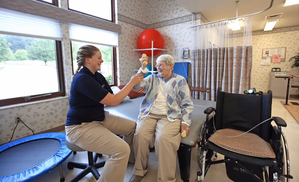 A female caregiver assists an elderly woman with arm exercises using small weights in a therapy room. The elderly woman is seated on a padded table next to a wheelchair. The room has large windows, a small trampoline, and exercise equipment in the background.