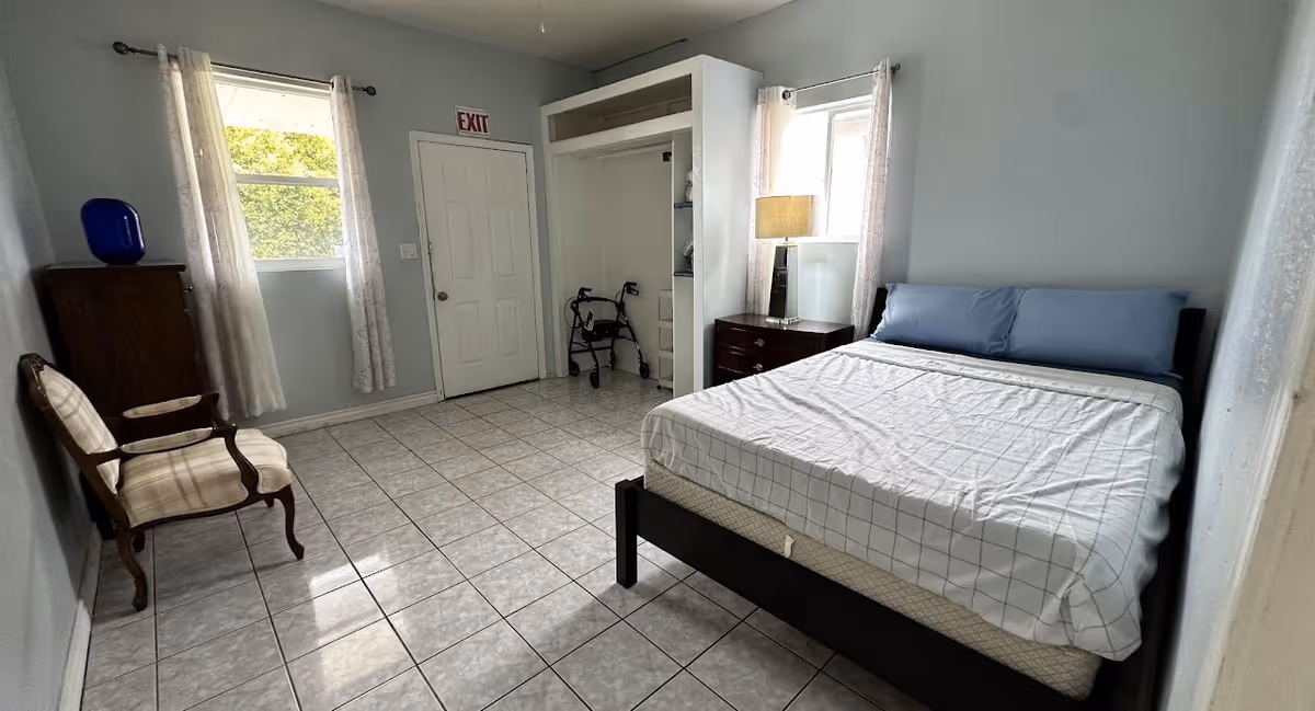 A simple bedroom with a bed covered in a white and gray checkered sheet and two blue pillows. There is a wooden nightstand with a lamp next to the bed, a wooden chair with a cushioned seat, a wooden cabinet with a blue vase on top, two windows with sheer curtains, a door with an exit sign above it, and an open closet containing a walker.
