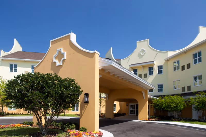 Exterior view of Discovery Village Trinity senior living facility showing a covered entrance with a curved roof, beige and light yellow walls, multiple windows, landscaped bushes, and colorful flower beds under a clear blue sky.