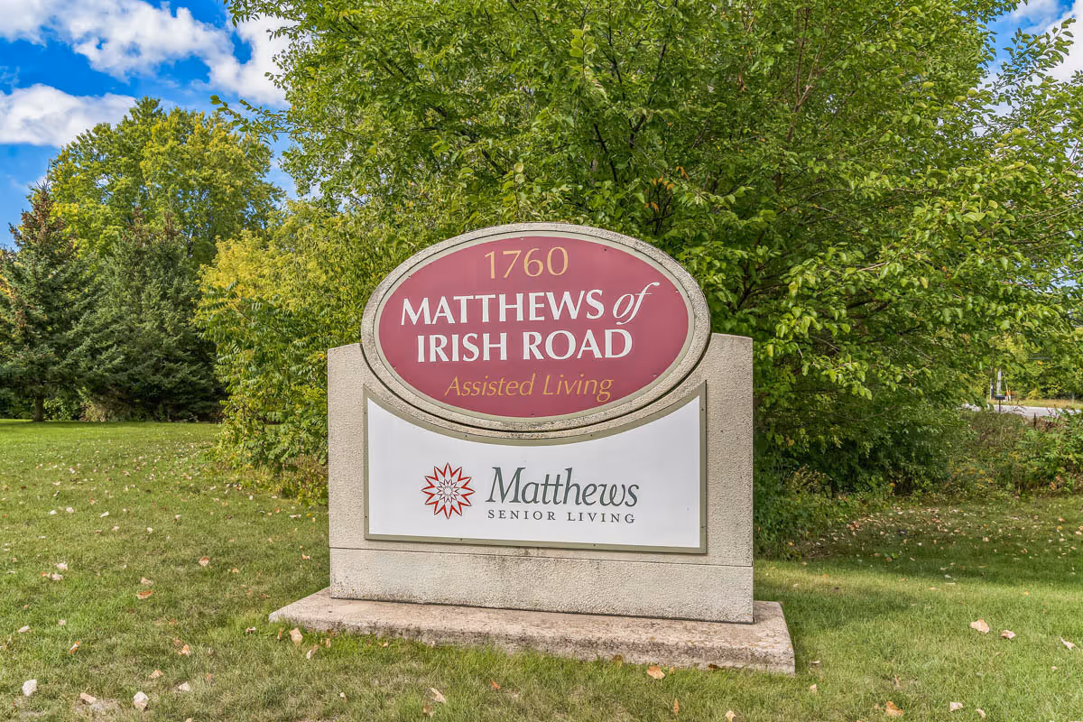 Outdoor image of a large stone sign on a grassy area with trees in the background. The sign reads '1760 Matthews of Irish Road Assisted Living' and 'Matthews Senior Living' with a red flower logo.