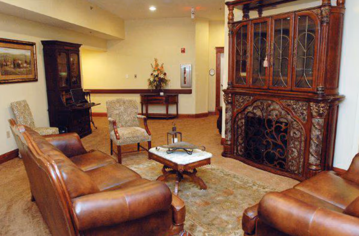 A cozy living room area with two brown leather sofas, two patterned armchairs, a small wooden coffee table with a decorative item, and an ornate wooden cabinet with intricate carvings and glass doors. The room has beige walls, carpeted flooring, a framed picture on the wall, and a small table with a flower arrangement in the background.