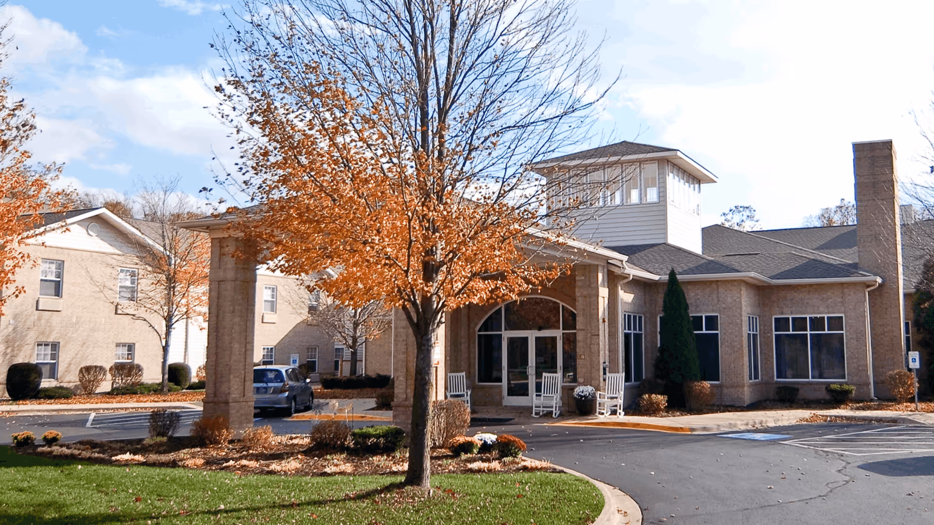 Exterior view of Cedarhurst Senior Living of Edison Lakes building with a covered entrance, rocking chairs on the porch, a tree with autumn leaves in the foreground, and a parking area with cars.