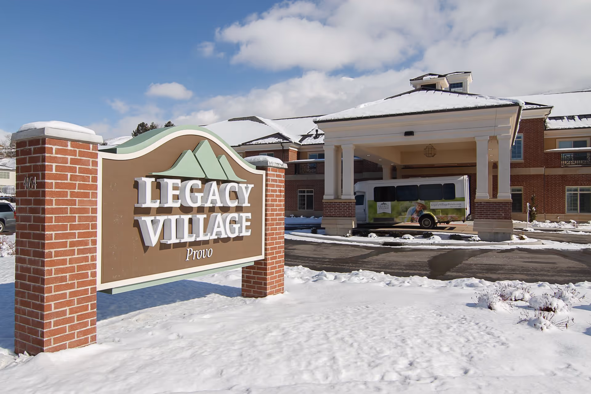 Snow-covered entrance of Legacy Village of Provo senior living facility with a large sign in the foreground and a covered drop-off area with a shuttle bus in the background under a partly cloudy sky.