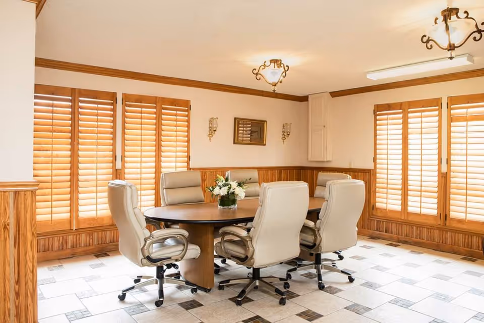 Round wooden table with six beige swivel chairs in a bright room with wooden shutters and a tiled floor.