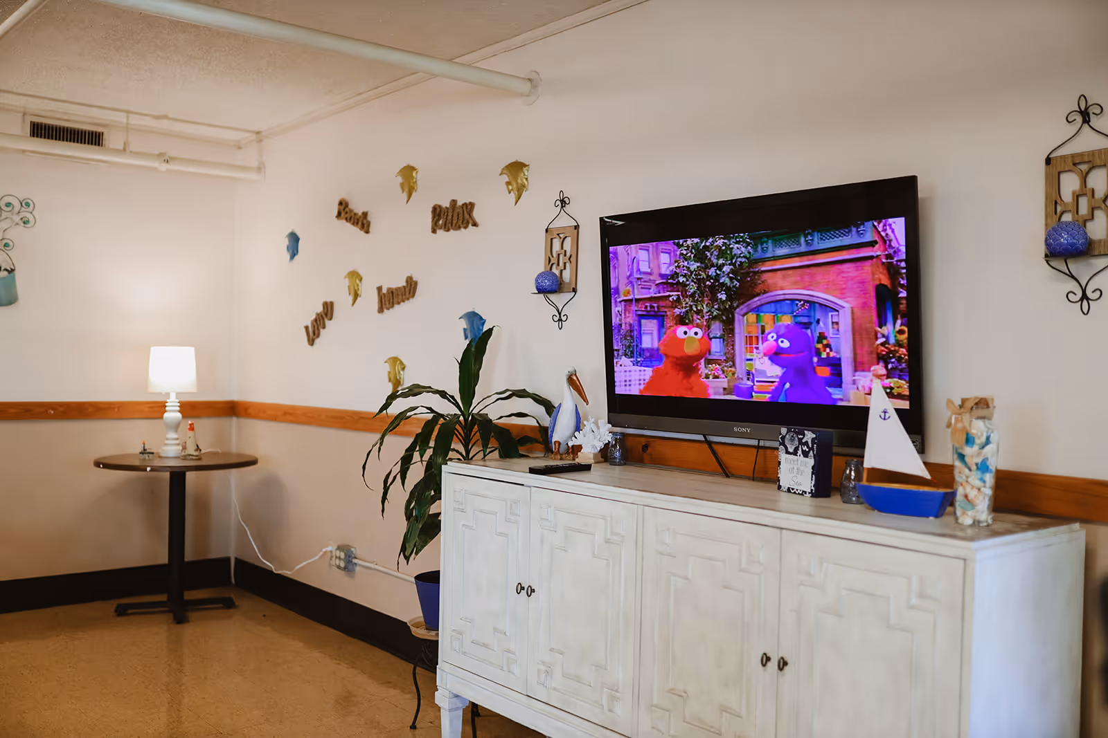 A cozy common area with a white cabinet holding decorative items including a small sailboat model and a pelican figurine. A flat-screen TV mounted on the wall displays a colorful children's show featuring Elmo and a purple character. The wall behind the TV has decorative words like 'Relax', 'Love', and 'Laugh' along with fish-shaped wall hangings. A small round table with a lamp is positioned in the corner, and a potted plant is placed next to the cabinet.