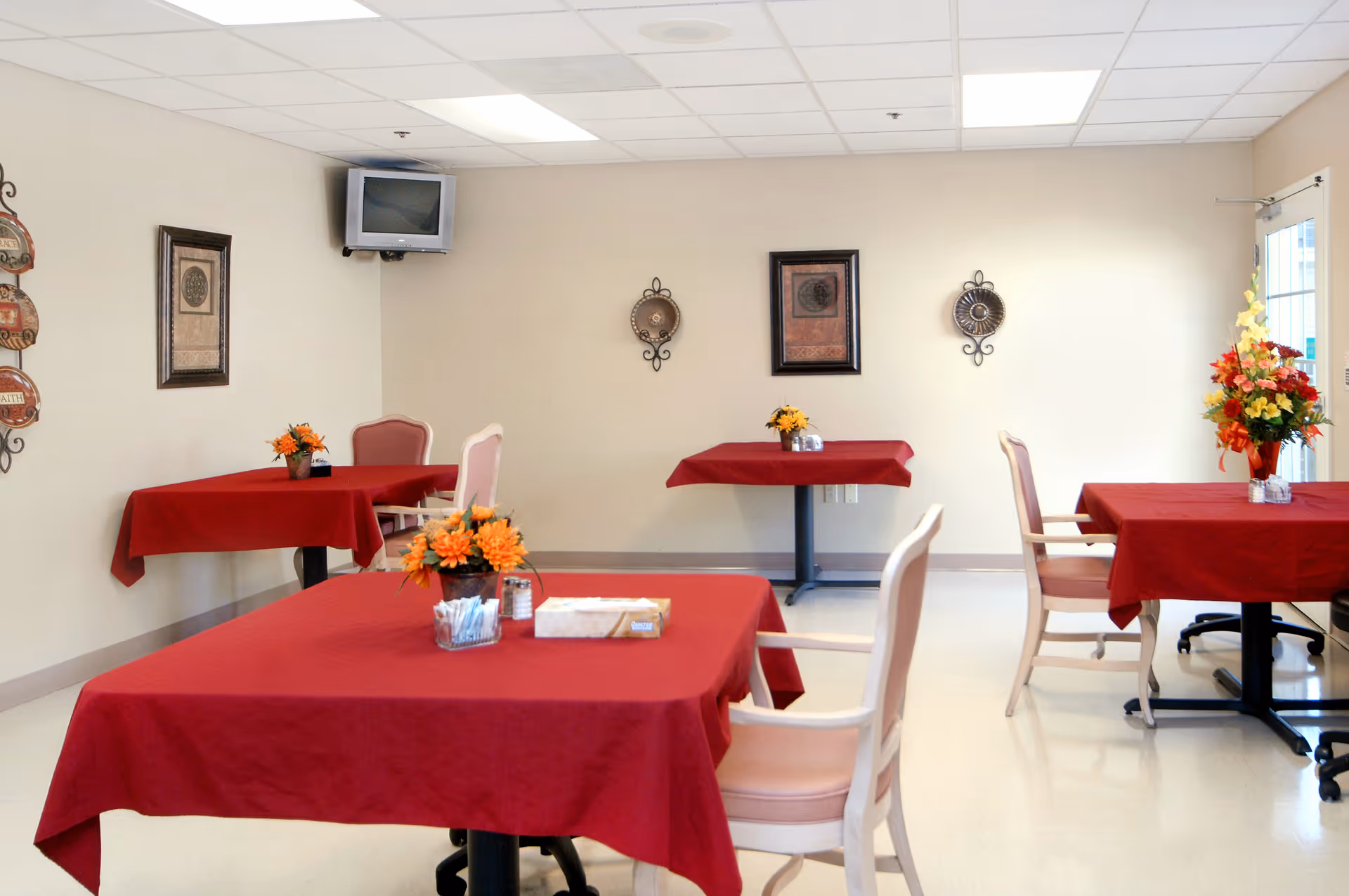 Dining area with tables covered in red tablecloths, chairs, small floral centerpieces and wall decor.