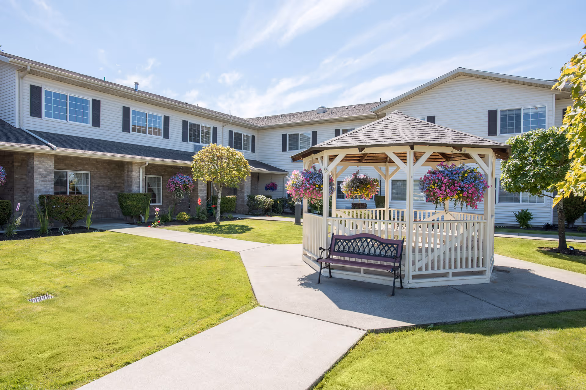 Sunny courtyard featuring a white gazebo with hanging flower baskets and a bench in front of a two-story senior living building.