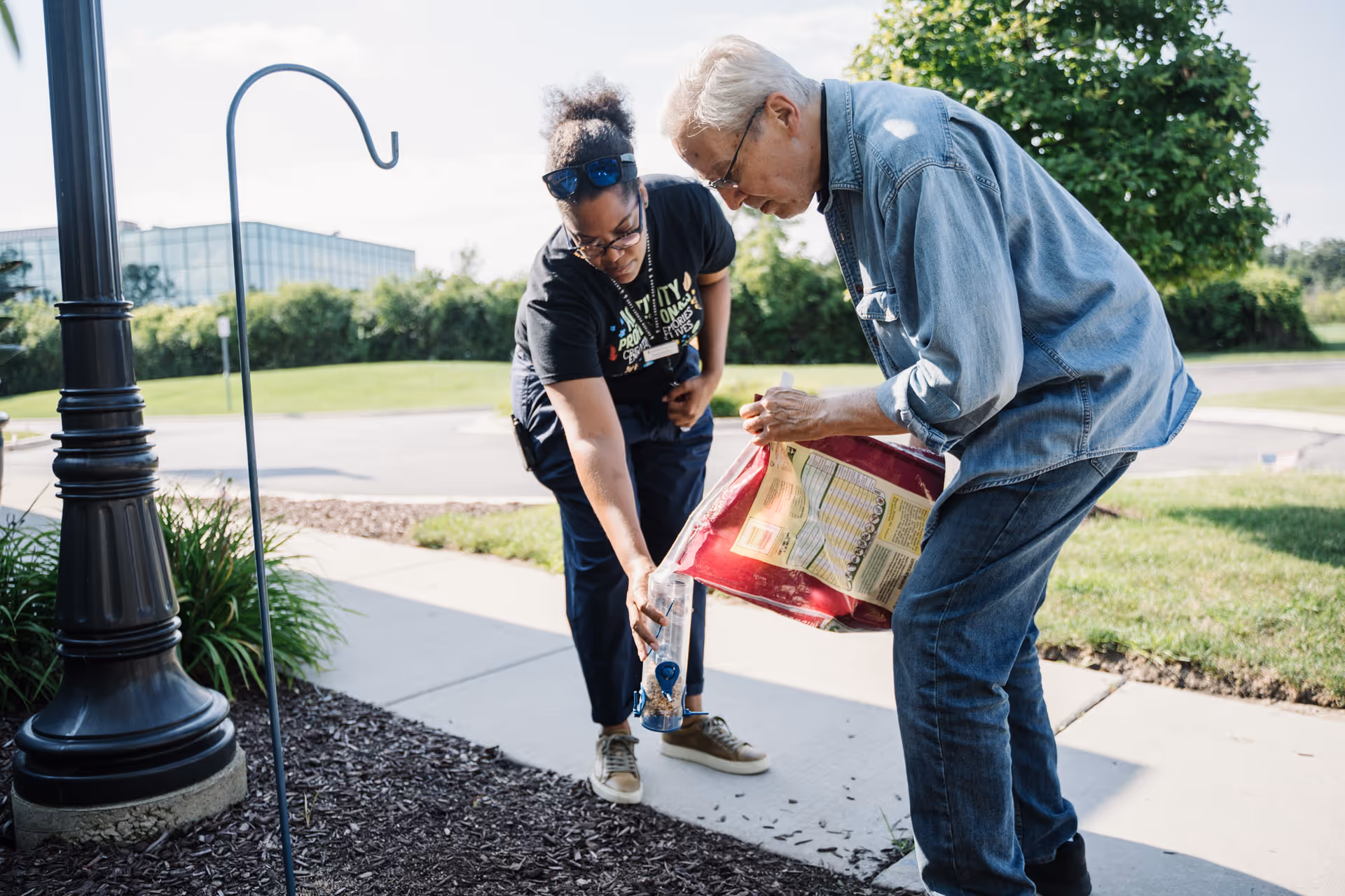 An elderly man and a woman outdoors near a sidewalk. The man is holding a large bag of birdseed and pouring it into a bird feeder held by the woman. They are surrounded by greenery and a lamppost is visible nearby.